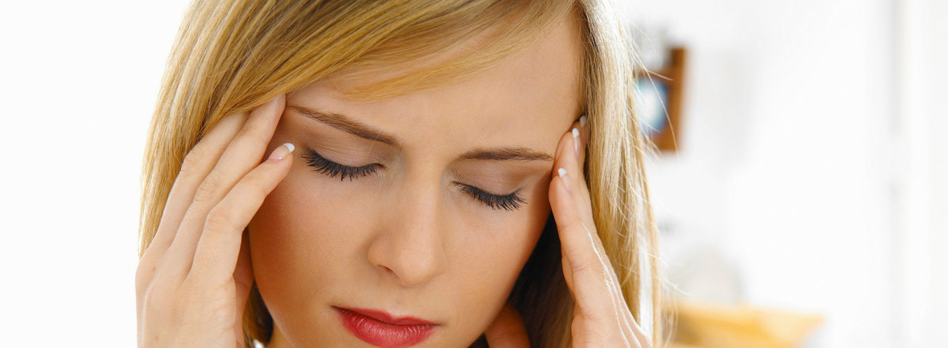 Junge blondhaarige Frau die sich aufgrund von Kopfschmerzen mit beiden Händen an die Schläfen packt