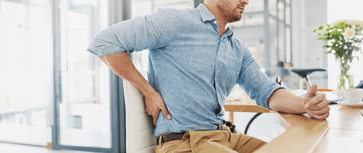 Mann mit blauem Hemd und brauner Hose sitz an einem Schreibtisch und hält sich Schmerzverzerrt den Rücken